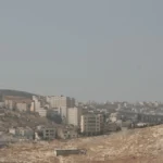 The Israeli separation wall cutting through residential areas and rocky terrain near Jerusalem, illustrating the severe spatial and developmental restrictions in the West Bank.