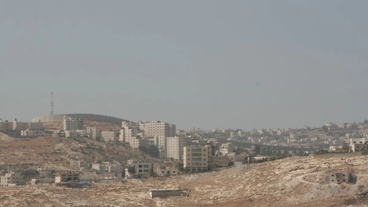 The Israeli separation wall cutting through residential areas and rocky terrain near Jerusalem, illustrating the severe spatial and developmental restrictions in the West Bank.