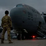 A U.S. Air Force Airman inspecting a C-17 Globemaster III cargo aircraft on the tarmac, representing the massive military supply chain and logistics network.
