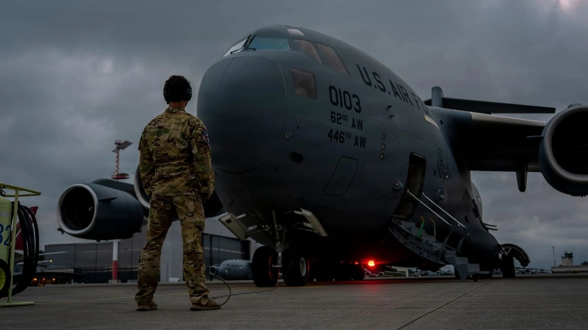 A U.S. Air Force Airman inspecting a C-17 Globemaster III cargo aircraft on the tarmac, representing the massive military supply chain and logistics network.