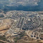Aerial view of an Israeli settlement in the occupied West Bank, showing dense concrete housing units strategically dividing the surrounding agricultural landscape.