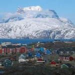 Colorful coastal houses of Nuuk's Nuussuaq district line the rocky Arctic shore, with the snow-covered Sermitsiaq mountain rising across the fjord in the background.